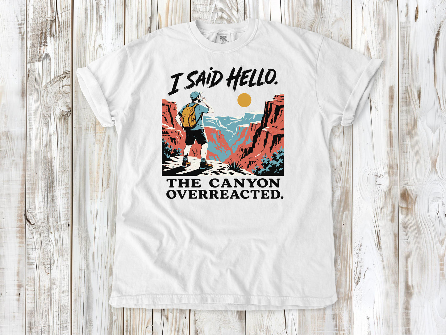 White garment-dyed tee with I Said Hello The Canyon Overreacted hiking graphic.