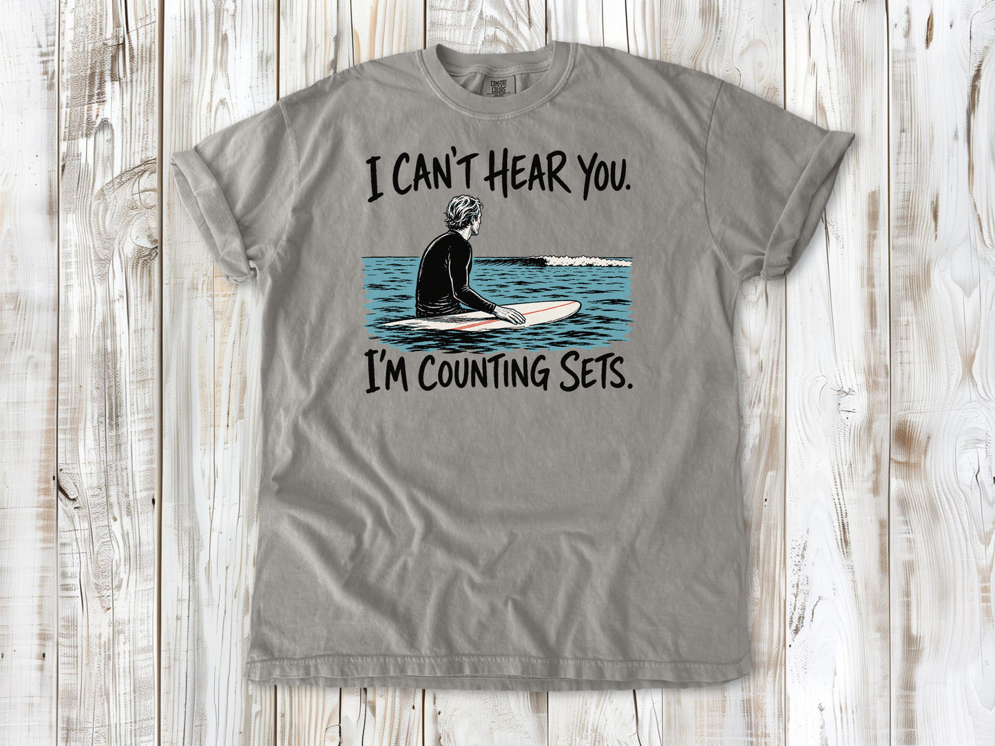 Khaki garment-dyed tee with I Can't Hear You I'm Counting Sets surfer graphic.