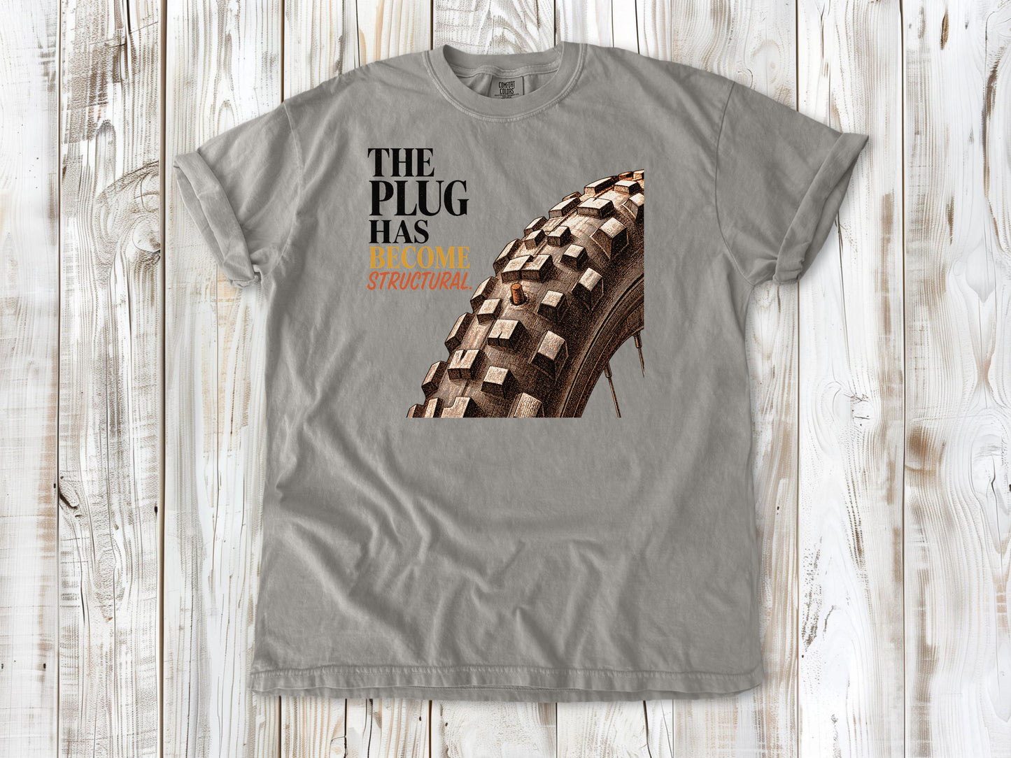 Khaki garment-dyed tee with The Plug Has Become Structural tubeless mountain bike tire graphic.