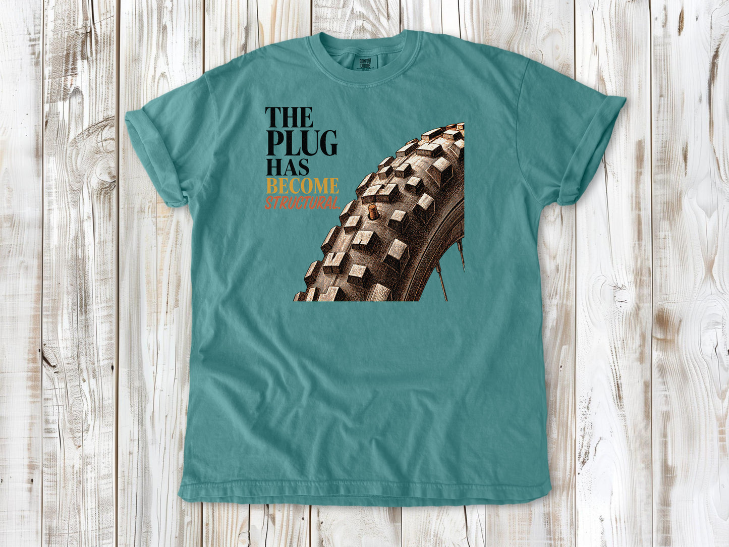 Mustard garment-dyed tee with The Plug Has Become Structural tubeless mountain bike tire graphic.