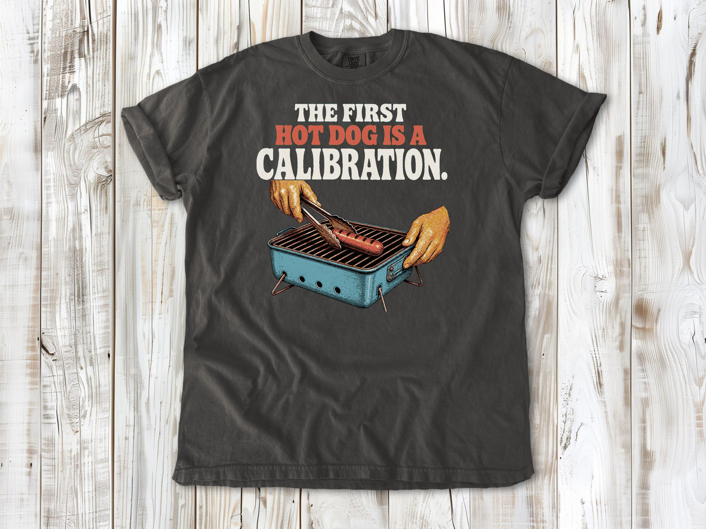Ivory garment-dyed tee with The First Hot Dog Is a Calibration beach grilling graphic.