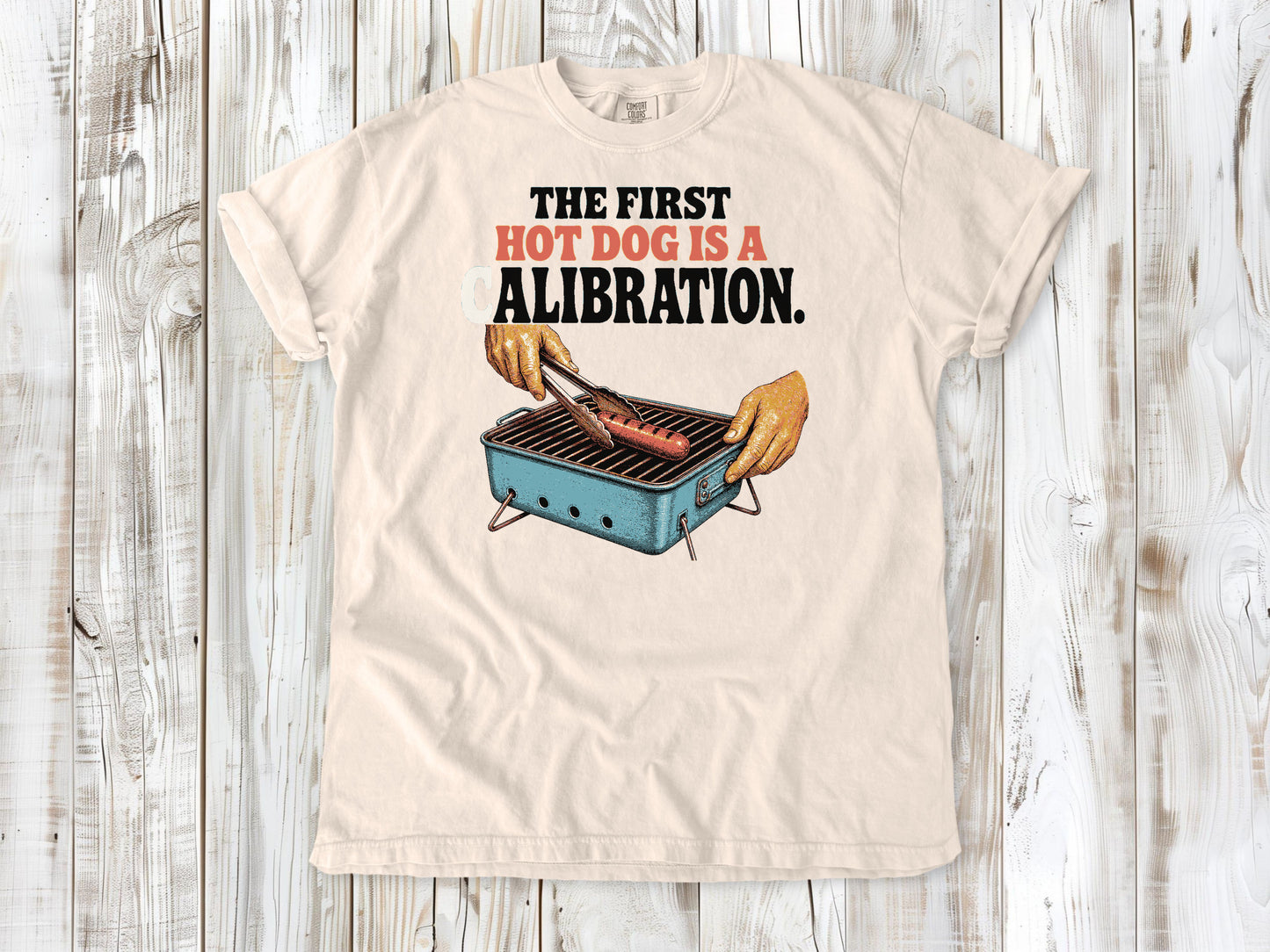 Butter garment-dyed tee with The First Hot Dog Is a Calibration beach grilling graphic.
