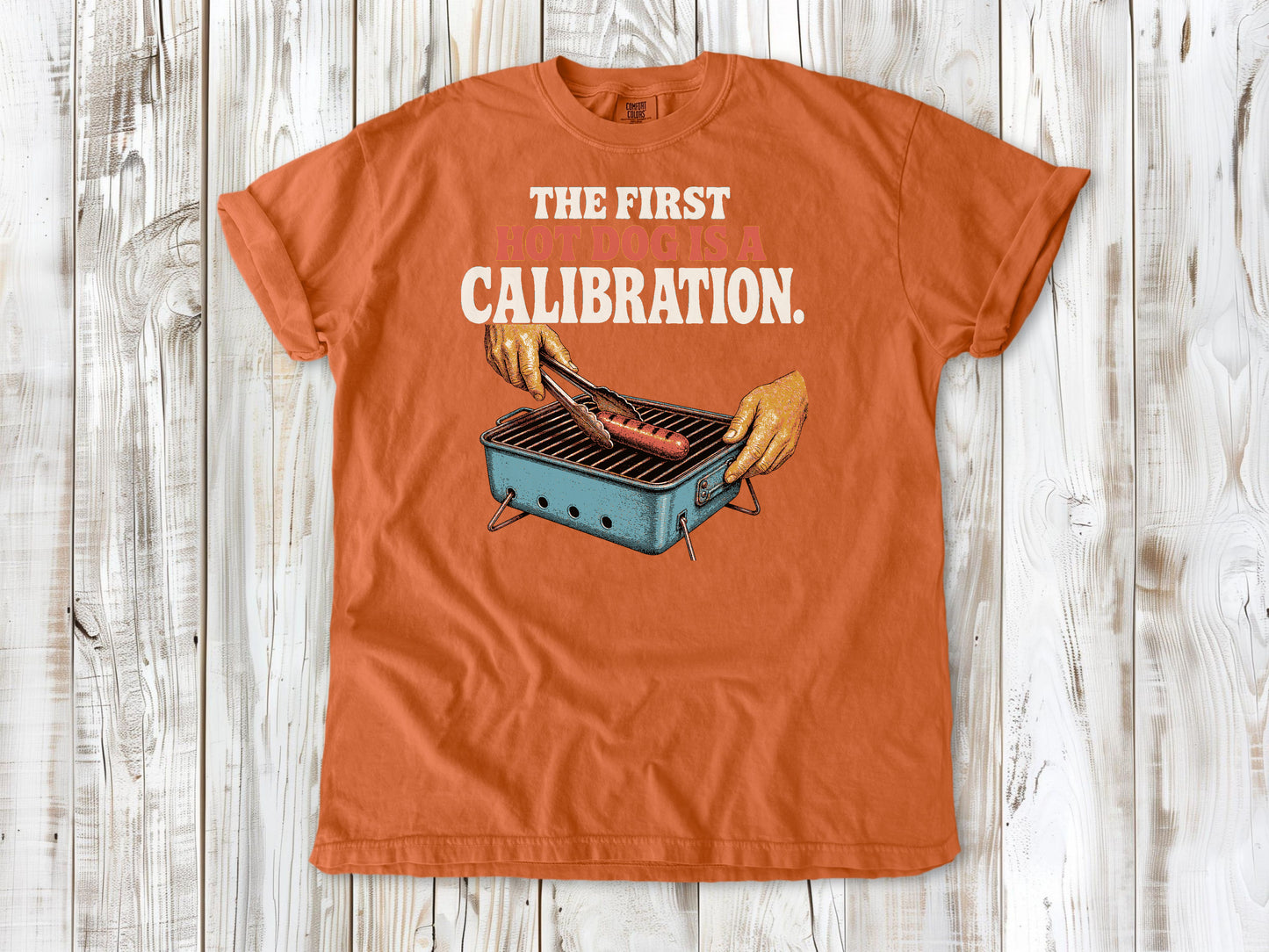 Seafoam garment-dyed tee with The First Hot Dog Is a Calibration beach grilling graphic.