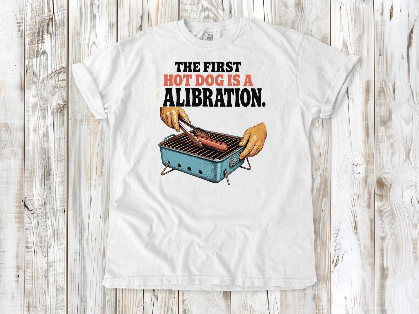 White garment-dyed tee with The First Hot Dog Is a Calibration beach grilling graphic.
