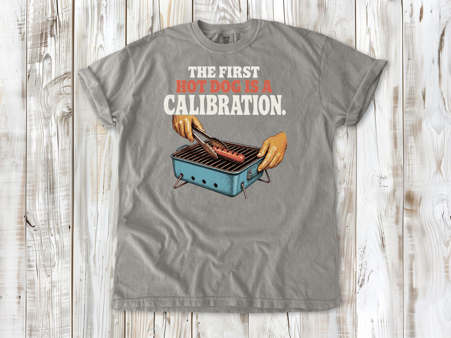 Khaki garment-dyed tee with The First Hot Dog Is a Calibration beach grilling graphic.