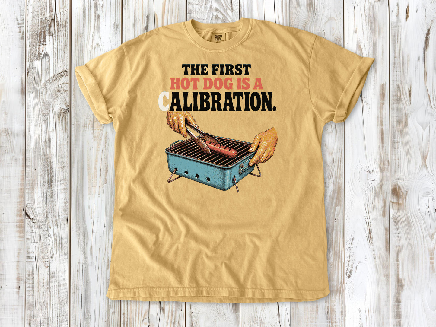 Burnt Orange garment-dyed tee with The First Hot Dog Is a Calibration beach grilling graphic.