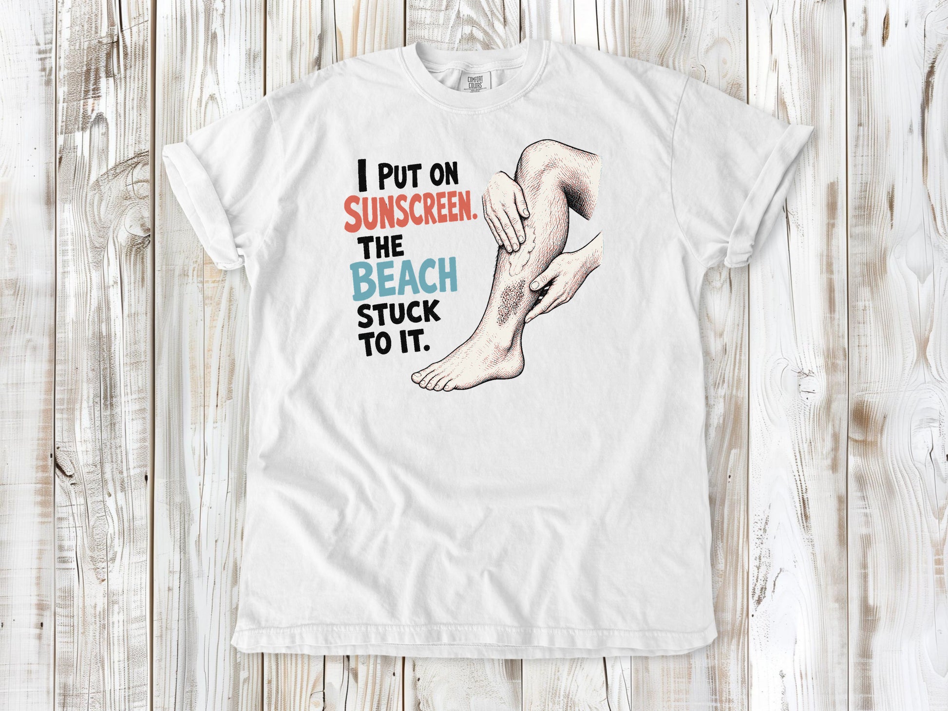 White garment-dyed tee with I Put on Sunscreen The Beach Stuck to It graphic.