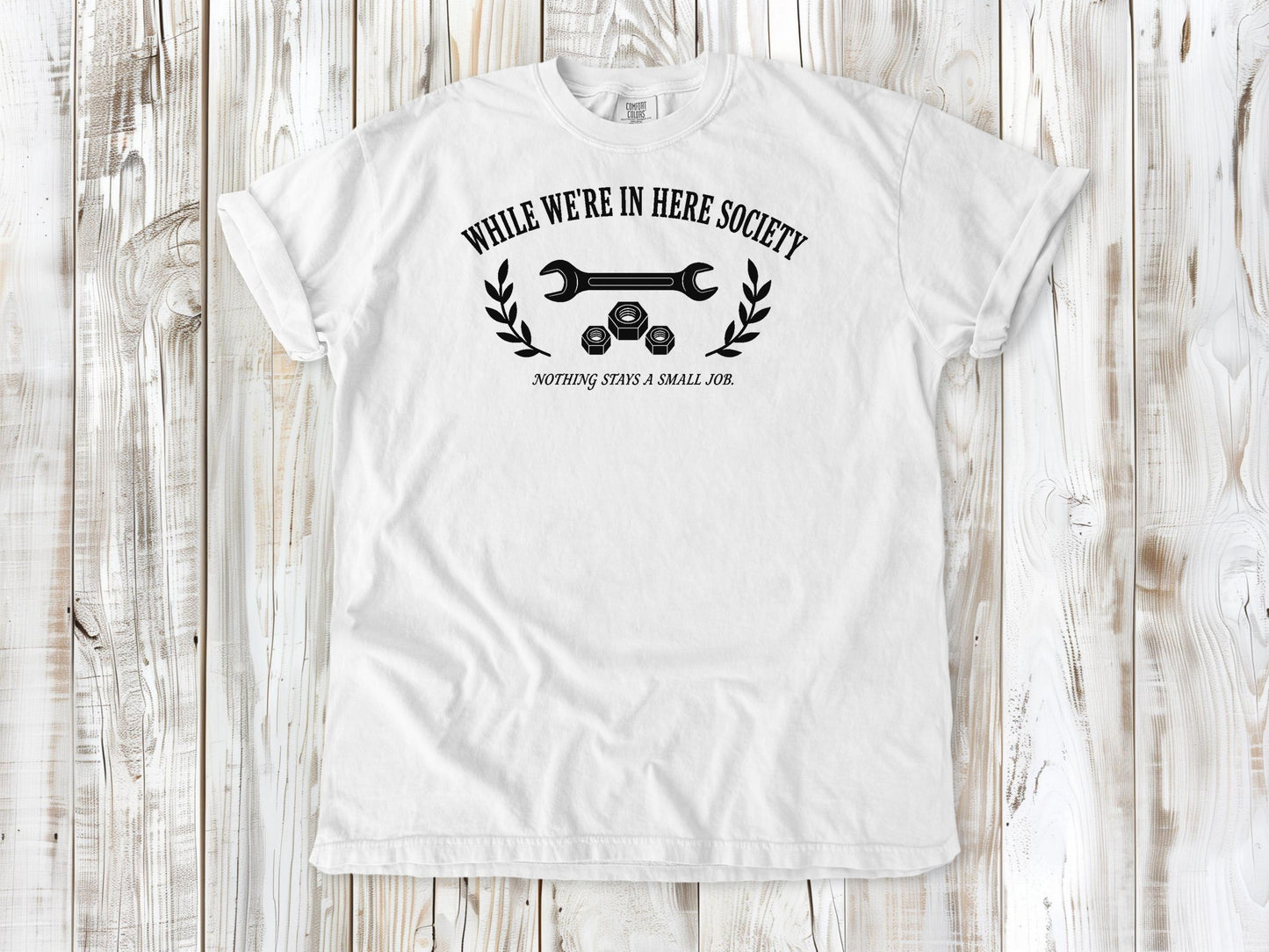 White garment-dyed tee with While We're in Here Society garage humor graphic.