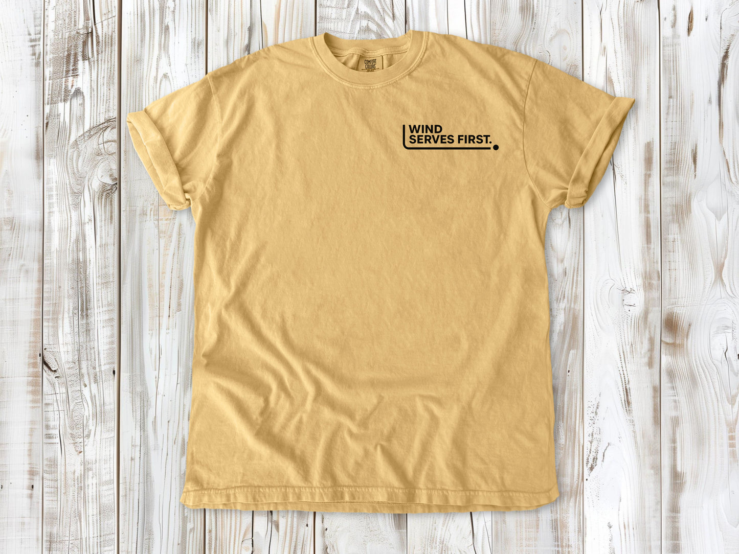 Mustard garment-dyed tee with Wind Serves First small left-chest beach volleyball graphic.