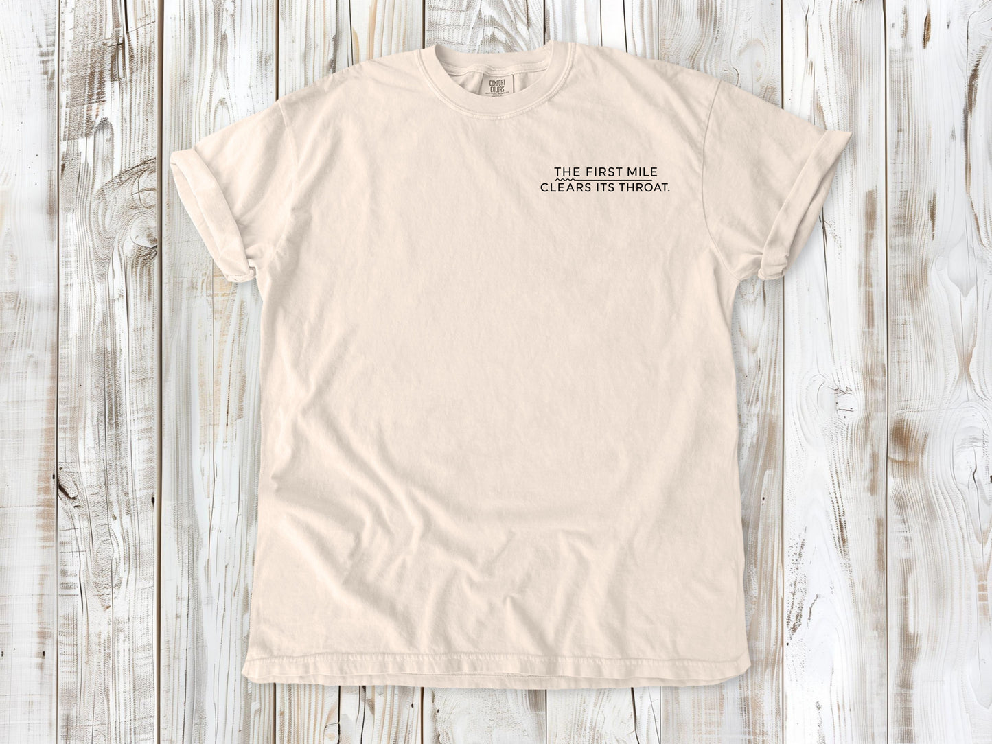 Ivory garment-dyed tee with The First Mile Clears Its Throat small left-chest running graphic.