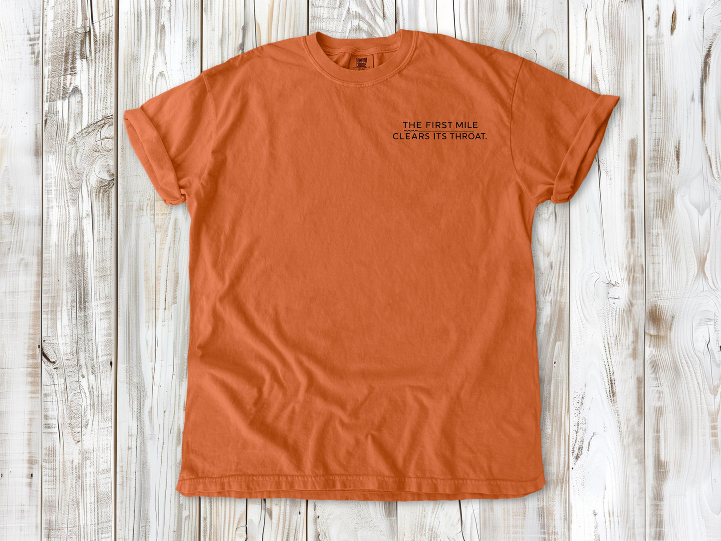 Burnt Orange garment-dyed tee with The First Mile Clears Its Throat small left-chest running graphic.