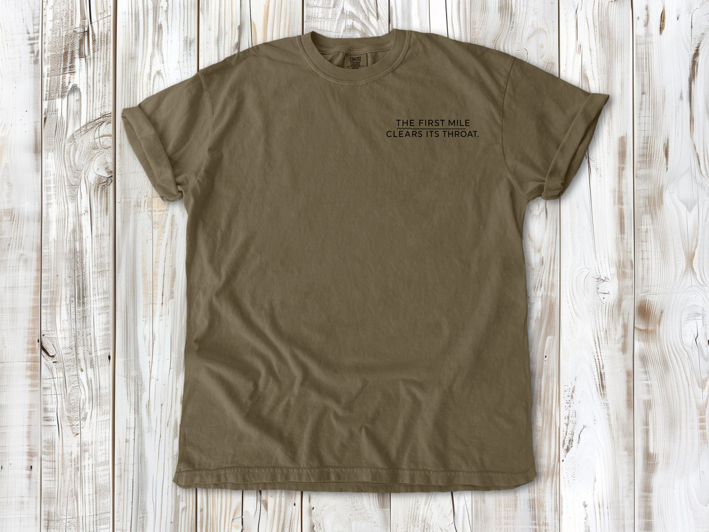Khaki garment-dyed tee with The First Mile Clears Its Throat small left-chest running graphic.