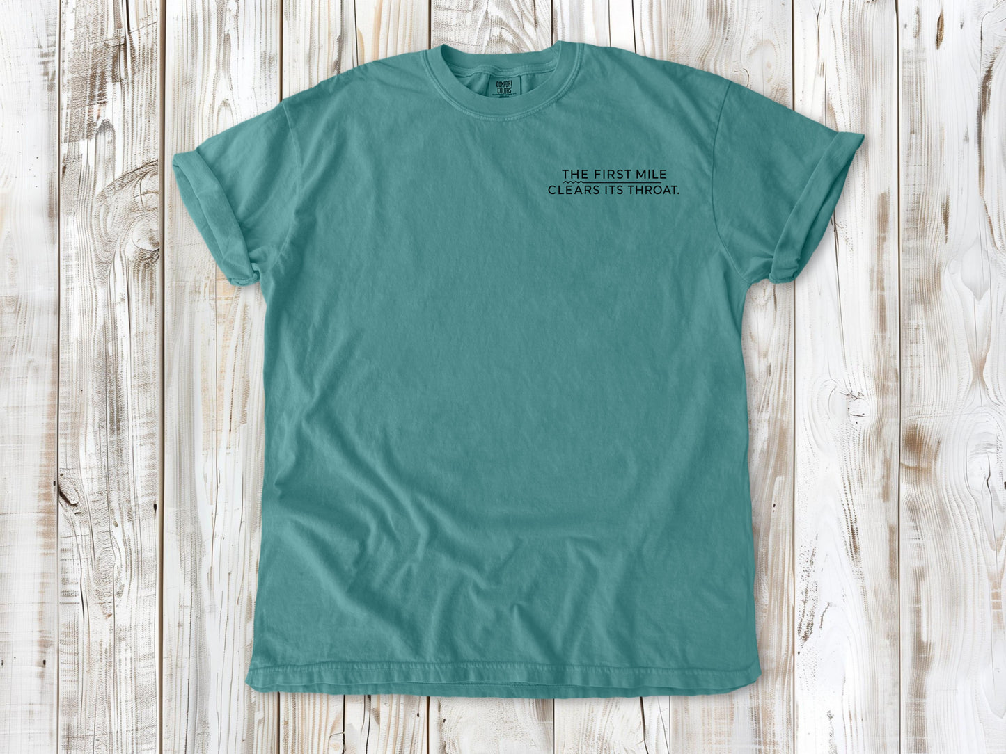 Seafoam garment-dyed tee with The First Mile Clears Its Throat small left-chest running graphic.