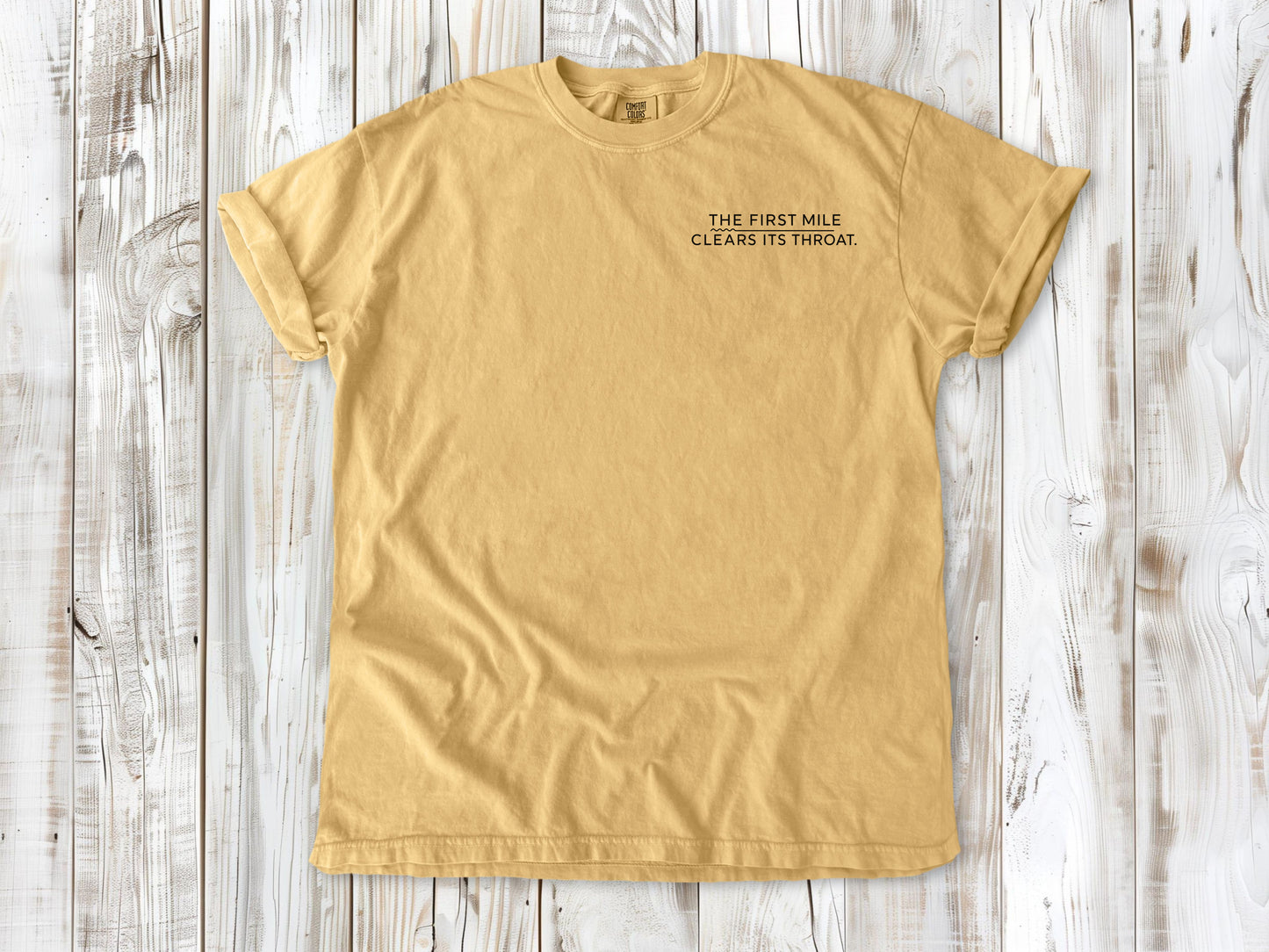 Mustard garment-dyed tee with The First Mile Clears Its Throat small left-chest running graphic.