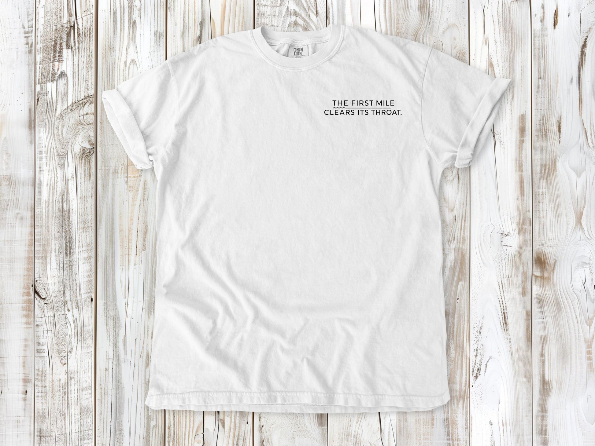 White garment-dyed tee with The First Mile Clears Its Throat small left-chest running graphic.