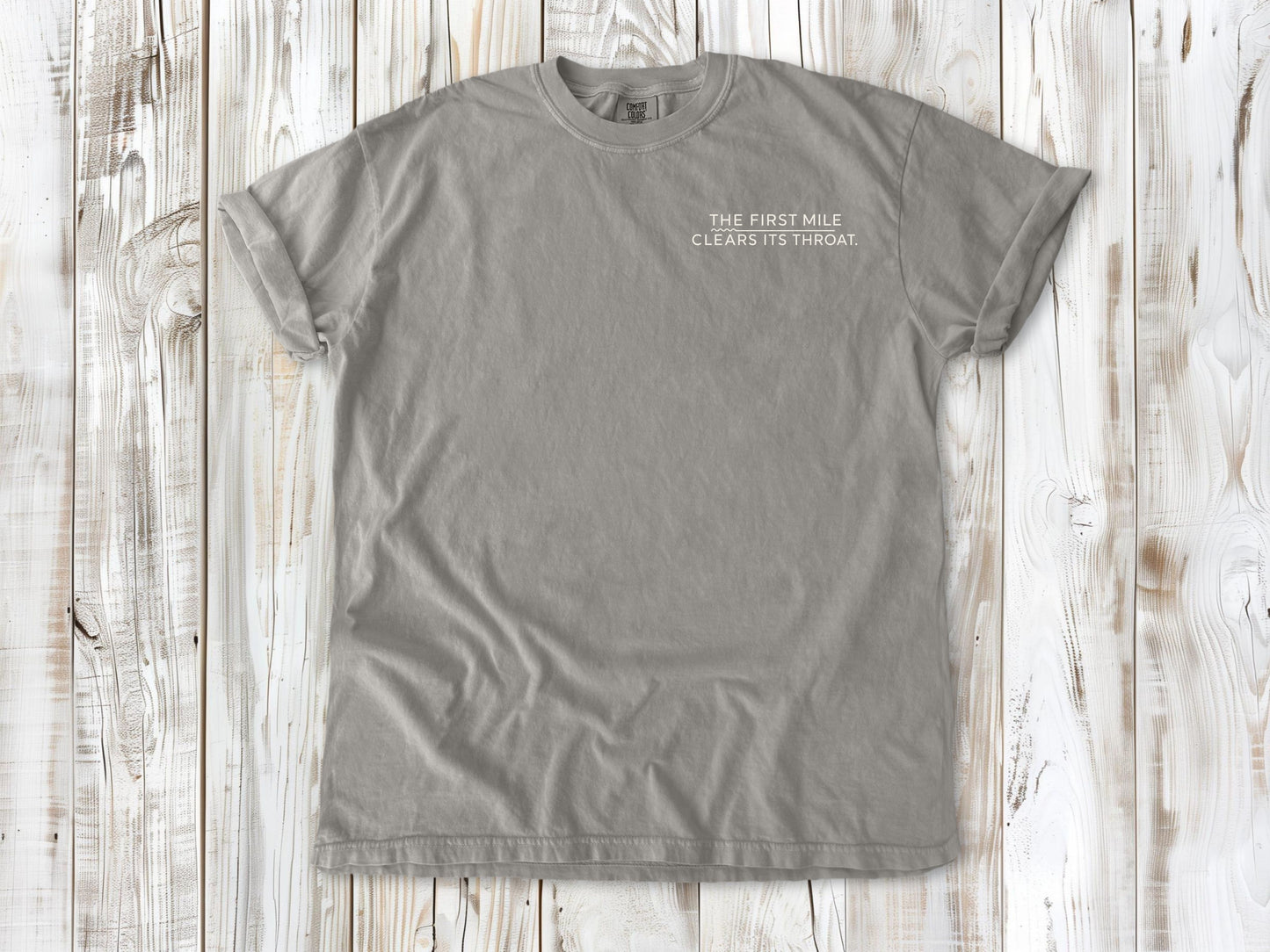 Grey garment-dyed tee with The First Mile Clears Its Throat small left-chest running graphic.
