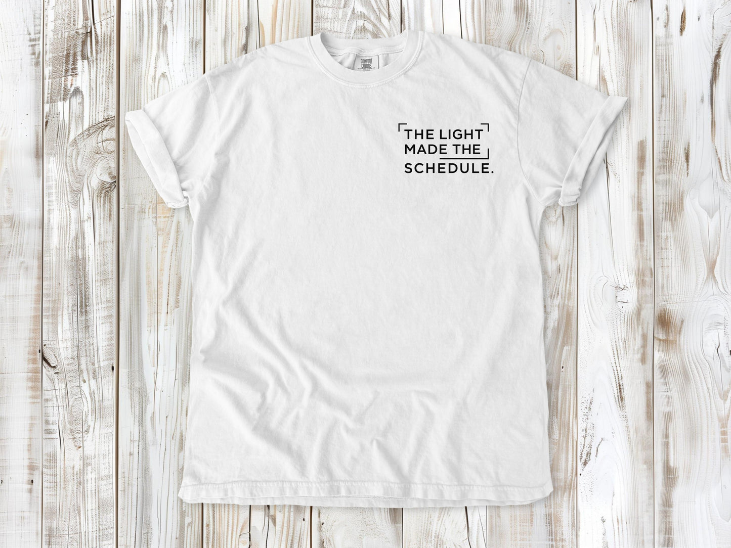 White garment-dyed tee with The Light Made the Schedule small left-chest photography graphic.