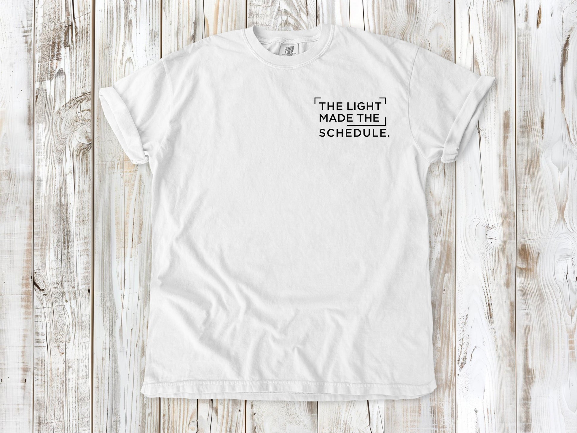 White garment-dyed tee with The Light Made the Schedule small left-chest photography graphic.