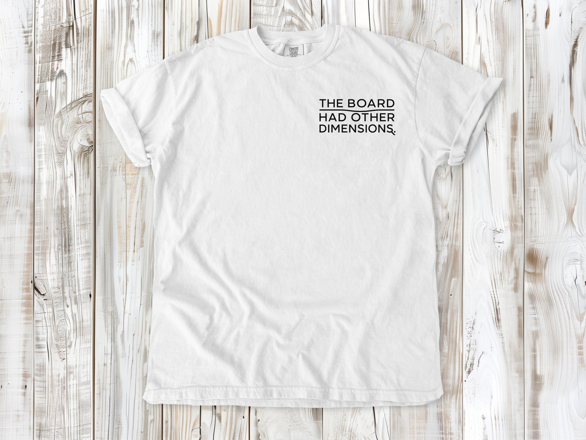 White garment-dyed tee with The Board Had Other Dimensions small left-chest woodworking graphic.