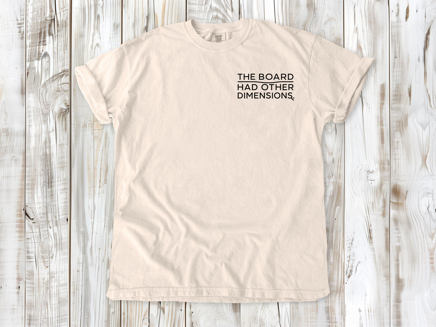 Ivory garment-dyed tee with The Board Had Other Dimensions small left-chest woodworking graphic.