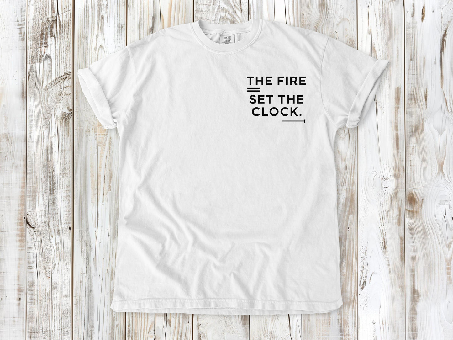 White garment-dyed tee with The Fire Set the Clock small left-chest camping graphic.