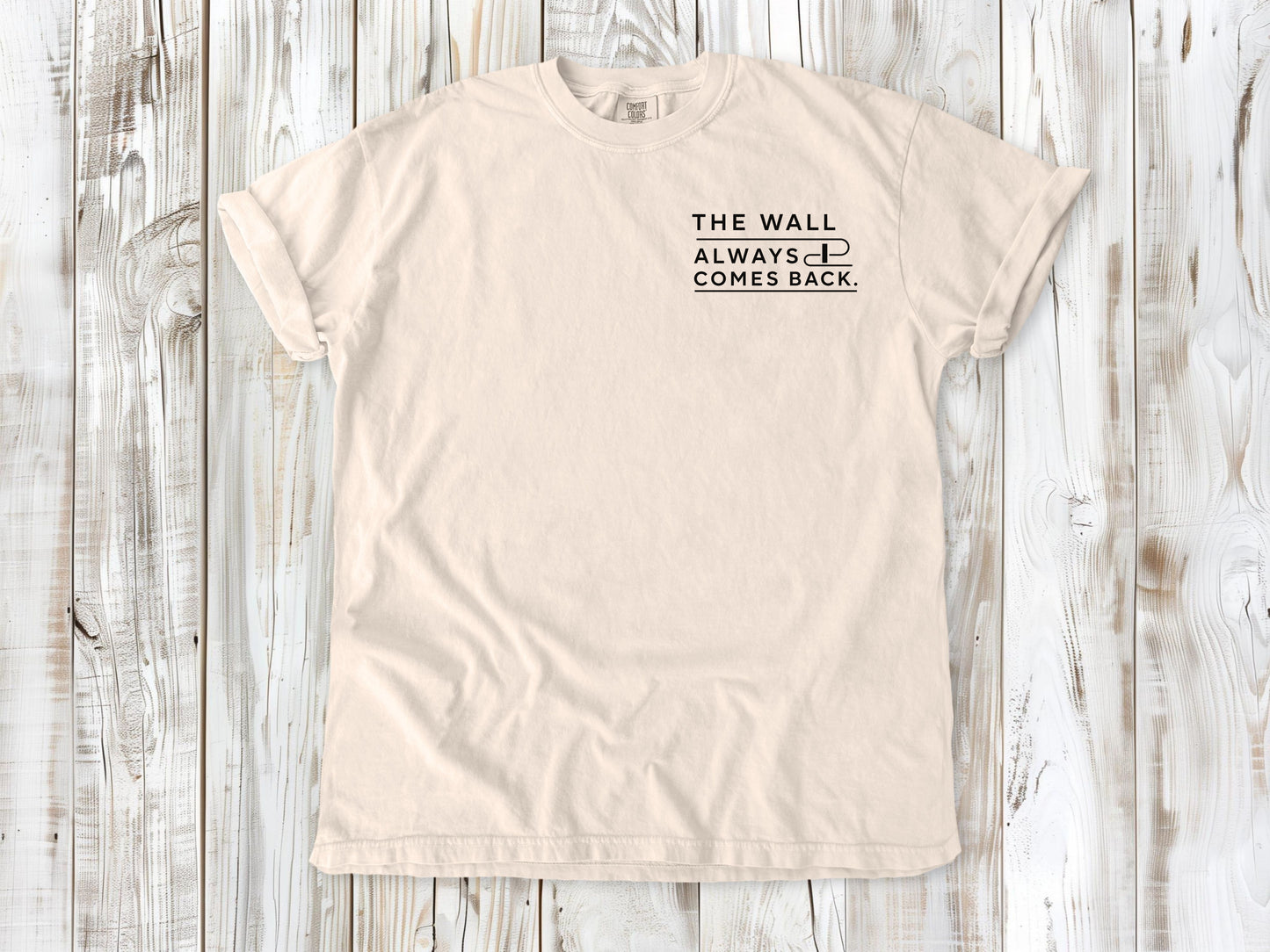 Ivory garment-dyed tee with The Wall Always Comes Back small left-chest swimming graphic.